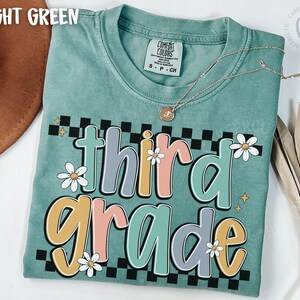 Op de afbeelding: Lichtgroen T-shirt met de woorden "third grade" in een kleurrijk, retro lettertype. Het ontwerp bevat madeliefjes en een zwart-wit geblokt patroon. Een gouden ketting is over het shirt gedrapeerd.