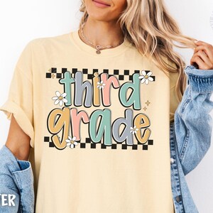 Op de afbeelding: Boterkleurig T-shirt met de woorden "third grade" in een kleurrijk, retro lettertype. De tekst staat tegen een zwart-witte geblokte achtergrond, met madeliefjesaccenten. Het shirt heeft opgerolde mouwen.