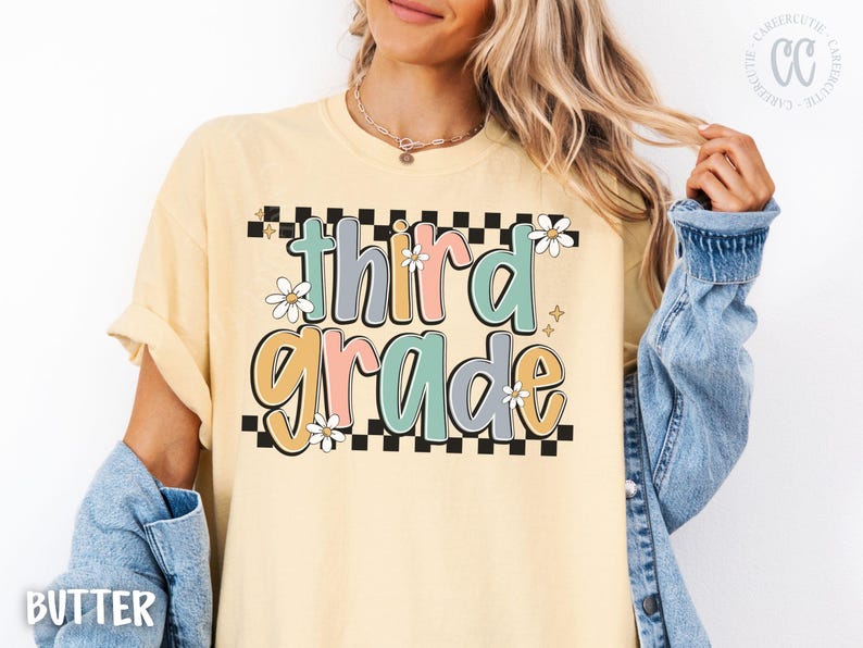 Op de afbeelding: Boterkleurig T-shirt met de woorden "third grade" in een kleurrijk, retro lettertype. De tekst staat tegen een zwart-witte geblokte achtergrond, met madeliefjesaccenten. Het shirt heeft opgerolde mouwen.