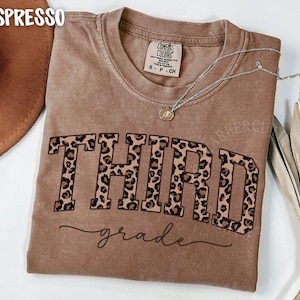 May include: Espresso-colored t-shirt with the word "THIRD" in large, leopard print letters and the word "grade" in cursive below. A silver necklace with a small pendant rests on the shirt. The shirt is a Comfort Colors brand.