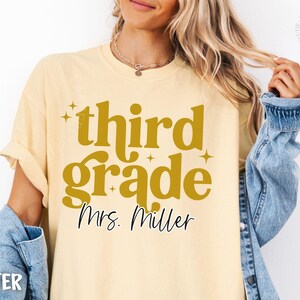May include: Pale yellow t-shirt featuring the text "third grade Mrs. Miller" in gold. The shirt has a casual style and is paired with a denim jacket. The text is in a retro font.