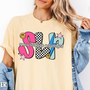 May include: Butter-colored t-shirt with a colorful graphic that says "SLP Speech Language Pathologist". The design includes a pink "S", a checkered "L", and a blue "P" with stars and smiley faces.
