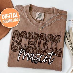 K&ouml;nnte beinhalten: Braunes T-Shirt mit dem Schriftzug "SCHOOL MASCOT" in Leopardenmuster. Das Shirt hat einen Vintage-Look. Eine silberne Halskette mit Anh&auml;nger liegt auf dem Shirt. Ein orangefarbener Kreis mit "DIGITALER DOWNLOAD" befindet sich oben links.