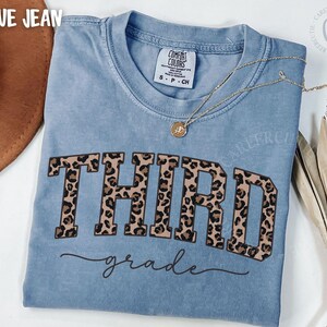May include: Blue jean Comfort Colors t-shirt with the word "THIRD" in leopard print block letters and "grade" in cursive. A gold necklace with a pendant is draped on the shirt. A brown hat is visible in the upper left corner.
