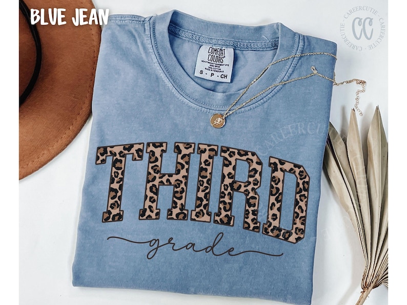 May include: Blue jean Comfort Colors t-shirt with the word "THIRD" in leopard print block letters and "grade" in cursive. A gold necklace with a pendant is draped on the shirt. A brown hat is visible in the upper left corner.