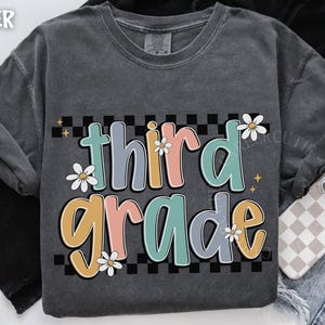 Op de afbeelding: Donkergrijze sweatshirt met de woorden "third grade" in kleurrijke, bubbelletters. De tekst staat tegen een zwart-wit geblokte achtergrond en is versierd met madeliefjes. Een casual, comfortabele stijl.