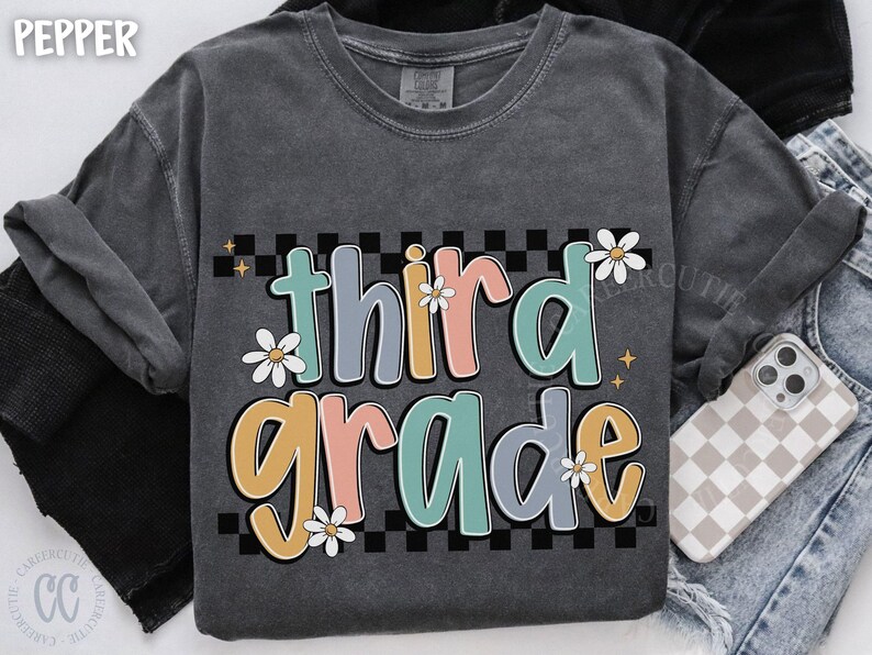 Op de afbeelding: Donkergrijze sweatshirt met de woorden "third grade" in kleurrijke, bubbelletters. De tekst staat tegen een zwart-wit geblokte achtergrond en is versierd met madeliefjes. Een casual, comfortabele stijl.