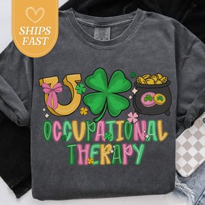 May include: Dark gray t-shirt with a colorful graphic design. The design includes a horseshoe with a pink bow, a green four-leaf clover, and a pot of gold coins. The text "OCCUPATIONAL THERAPY" is in green and pink.
