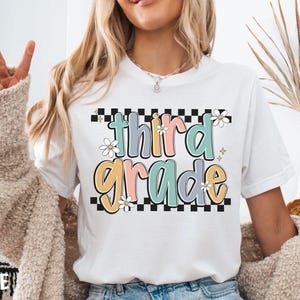 Op de afbeelding: Wit T-shirt met een kleurrijke "third grade" afbeelding. Het ontwerp heeft pastelkleuren, madeliefjes en een geblokte rand. Een casual T-shirt voor leerlingen of leraren.