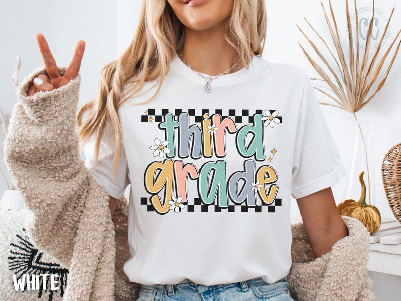 Op de afbeelding: Wit T-shirt met een kleurrijke "third grade" afbeelding. Het ontwerp heeft pastelkleuren, madeliefjes en een geblokte rand. Een casual T-shirt voor leerlingen of leraren.