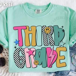 May include: A light green t-shirt with the words "THIRD GRADE" in a colorful, retro font. The design includes stars, a lightning bolt, and smiley faces. Sunglasses and bracelets are visible.