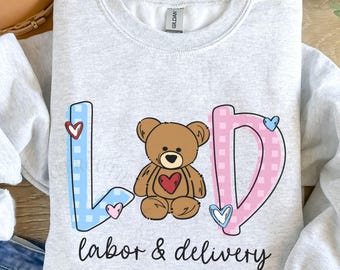 Op de afbeelding: Asgrijze sweatshirt met de tekst "L&D labor & delivery". De letters L en D zijn in blauwe en roze geruite patronen, met een bruine teddybeer in het midden. Kleine hartjes zijn verspreid rond de letters.