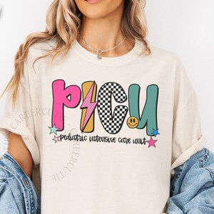 May include: Ivory-colored t-shirt with a colorful "PICU" graphic. The design includes a pink "P", a yellow lightning bolt, a black and white checkered "C", and a teal "U". Below the graphic is the text "pediatric intensive care unit".