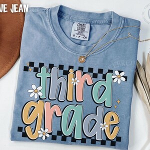 Op de afbeelding: Blauw Comfort Colors T-shirt met een kleurrijke "third grade" graphic. Het ontwerp heeft madeliefjes, een dambordpatroon en pastelkleuren. Een gouden ketting is over het shirt gedrapeerd.
