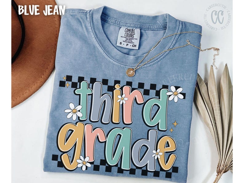 Op de afbeelding: Blauw Comfort Colors T-shirt met een kleurrijke "third grade" graphic. Het ontwerp heeft madeliefjes, een dambordpatroon en pastelkleuren. Een gouden ketting is over het shirt gedrapeerd.