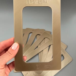 May include: A collection of tan-coloured rectangular frames with rounded corners. The top frame has the word "BRONZORA" embossed at the top. Additional frames are visible in the background. Measurements are printed on the frames.