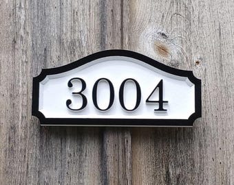 May include: A rectangular address plaque with the number "3004" in black, centered on a white background. The plaque has a decorative black border and is mounted on a weathered wooden surface.