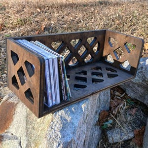 May include: A dark brown wooden CD storage shelf with a geometric lattice design. The shelf is filled with a stack of CDs and is sitting on a rock.