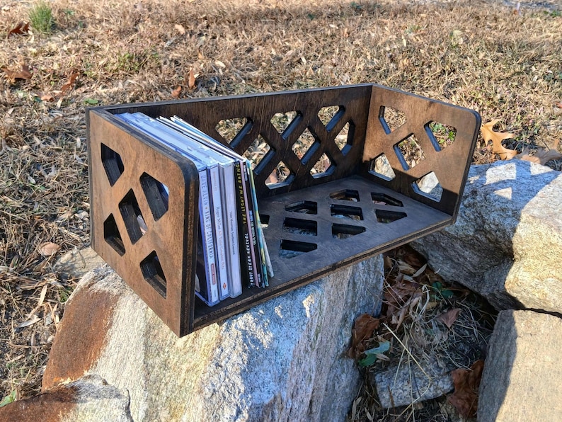 May include: A dark brown wooden CD storage shelf with a geometric lattice design. The shelf is filled with a stack of CDs and is sitting on a rock.