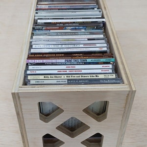 May include: A wooden crate with a grid pattern on the front, filled with compact discs. The CDs are stacked vertically and feature various artists and album titles, including "Meat Loaf", "Old Crow Medicine Show", "John Prine", and "Billy Joe Shaver".