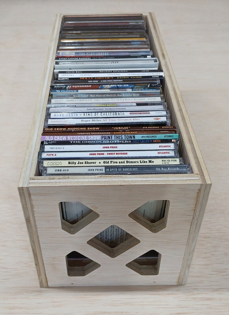 May include: A wooden crate with a grid pattern on the front, filled with compact discs. The CDs are stacked vertically and feature various artists and album titles, including "Meat Loaf", "Old Crow Medicine Show", "John Prine", and "Billy Joe Shaver".
