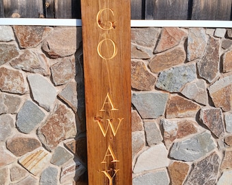 May include: A rectangular wooden sign with the words "GO AWAY" carved into the surface. The sign is made of a dark brown wood and the letters are a lighter shade of brown. The sign is displayed outdoors.
