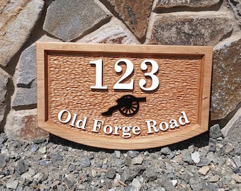 May include: A wooden sign with a carved, textured surface. The sign features the house number "123" in white, a black silhouette of a cannon, and the text "Old Forge Road" in white.