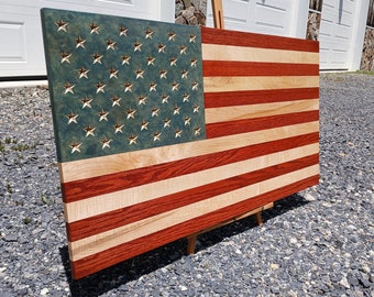 May include: A wooden American flag with red, white, and blue stripes. The stars are carved into the blue section of the flag. The flag is displayed on an easel.
