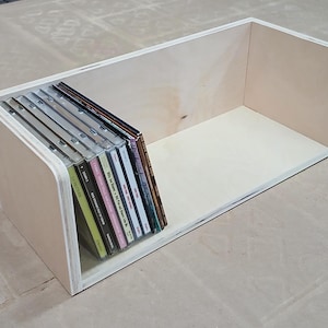 May include: A wooden box with a rounded front edge, designed to hold and display a collection of CDs. The box is made of light-coloured wood and has a natural finish. The CDs are arranged vertically in the box, with their spines facing outward.