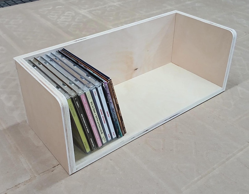 May include: A wooden box with a rounded front edge, designed to hold and display a collection of CDs. The box is made of light-coloured wood and has a natural finish. The CDs are arranged vertically in the box, with their spines facing outward.