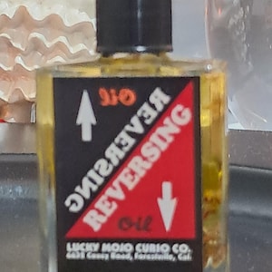 May include: A small glass bottle with a black cap filled with a yellow liquid. The label is black with red and white text that reads "REVERSING OIL" and "016" with an arrow pointing up and another arrow pointing down. The label also includes the text "LUCKY MOJO CURIO CO. 6631 Costy Road, Forestville, Cal."