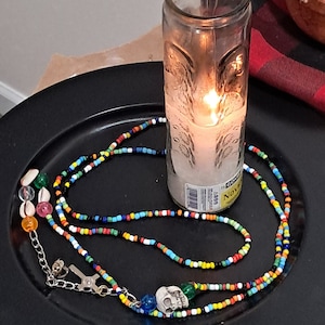 May include: A colorful beaded necklace with a skull pendant and various charms rests on a black plate. A lit candle in a glass jar sits nearby. The necklace features a variety of small, brightly colored beads.
