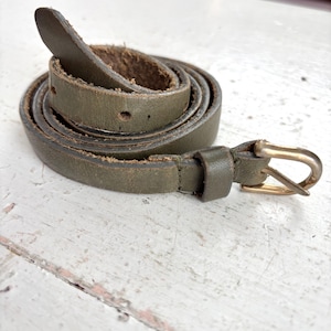 May include: Olive green leather belt with a gold-colored buckle. The belt is rolled up, showing the texture of the leather and the details of the buckle. The belt has several holes for adjustment.