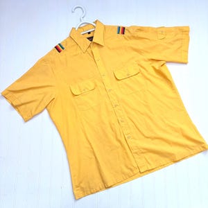 May include: Yellow short-sleeved shirt with two chest pockets and button-down front. The shoulders have a colorful striped detail. The shirt is hanging on a white hanger.