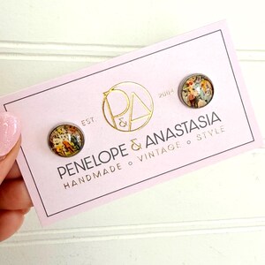 May include: Two round stud earrings featuring a colorful, detailed vintage-style illustration under a glass dome. The earrings are mounted on a light pink rectangular display card with the text PENELOPE & ANASTASIA HANDMADE VINTAGE STYLE.