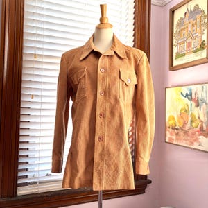 May include: A tan corduroy shirt with a button-up front, two chest pockets, and a pointed collar. The long-sleeved shirt is displayed on a mannequin. The shirt has a vintage style.