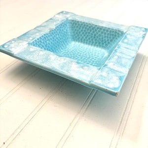 May include: A square, light blue ceramic dish with a textured interior and a raised, textured border. The dish has a slightly distressed appearance, with white accents on the edges.