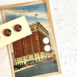 May include: A pair of round stud earrings with a vintage building design, displayed on a cream-colored card. The earrings are set against a vintage postcard of the Hotel Melbourne in St. Louis, Missouri.