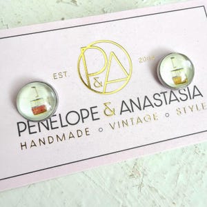 May include: Two round stud earrings featuring a small graphic of a red and yellow cable car on a light background. The earrings are displayed on a light pink card with the gold text P&A EST. 2004 PENELOPE & ANASTASIA HANDMADE VINTAGE STYLE.