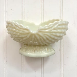 May include: Cream-colored, vintage-style glass bowl with a scalloped rim and textured, shell-like design. The bowl sits on a pedestal base with embossed detailing. A decorative piece for display or serving.