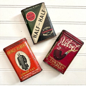 May include: Three vintage tobacco tins with colorful labels. The tins are red, green, and brown with gold lettering. The text on the tins reads "Prince Albert", "Half and Half", and "Velvet".