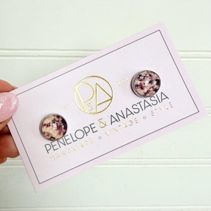 May include: A pair of round stud earrings featuring a mottled pattern of dark red, black, and cream colors. The earrings are attached to a rectangular white display card with the gold text Penelope & Anastasia, Handmade, Vintage, Style.
