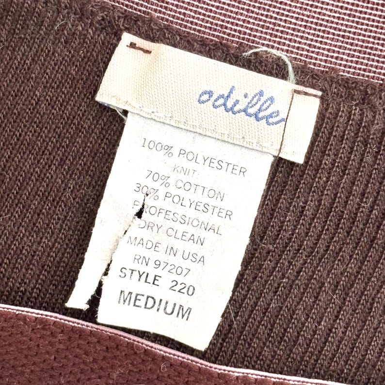 May include: Close-up of a brown knit garment with a tag. The tag reads "odille" in blue script, and lists fabric composition as 100% polyester, 70% cotton, and 30% polyester. The tag also indicates "MADE IN USA" and "MEDIUM" size.