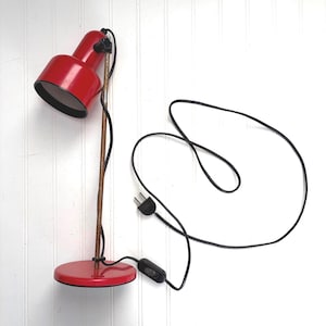 May include: A red desk lamp with a black power cord. The lamp has a round base and a cylindrical stem. The lamp head is also cylindrical and can be adjusted. The lamp is approximately 12 inches tall.