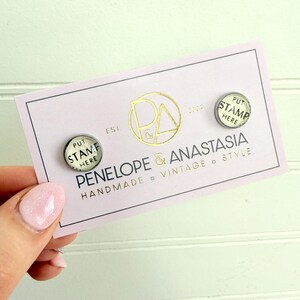 May include: A pair of round, silver-toned stud earrings with the text "PUT STAMP HERE" on a white background. The earrings are displayed on a pink card with gold lettering that reads "PENELOPE & ANASTASIA HANDMADE VINTAGE STYLE."