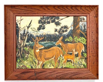 May include: A framed oil painting depicting a deer family in a forest landscape. The scene features a buck, a doe, and a fawn standing in tall green grass near a body of water, surrounded by pine trees, all encased in a rustic wooden frame.