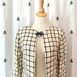 May include: A cream-colored jacket with a black and white plaid pattern. The jacket has a mandarin collar and a single button closure at the neck. The button is a dark blue knot.