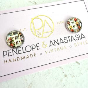 May include: Two round stud earrings featuring a floral pattern with white, green, and red tones, displayed on a white card with the text PENELOPE & ANASTASIA, EST. 2004, HANDMADE, VINTAGE, STYLE, and a gold circular logo with the letters P and A.