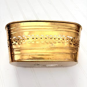 May include: A gold-colored, oval-shaped planter with a decorative, openwork band around the middle. The planter has a rounded rim and a flat, oval base. The metal has a shiny finish and shows some signs of wear.