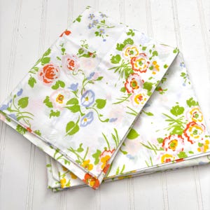 May include: White floral bed sheets with a colorful pattern of roses, morning glories, and other flowers in shades of orange, pink, yellow, blue, and green. The sheets are folded and stacked, showcasing the vibrant design.